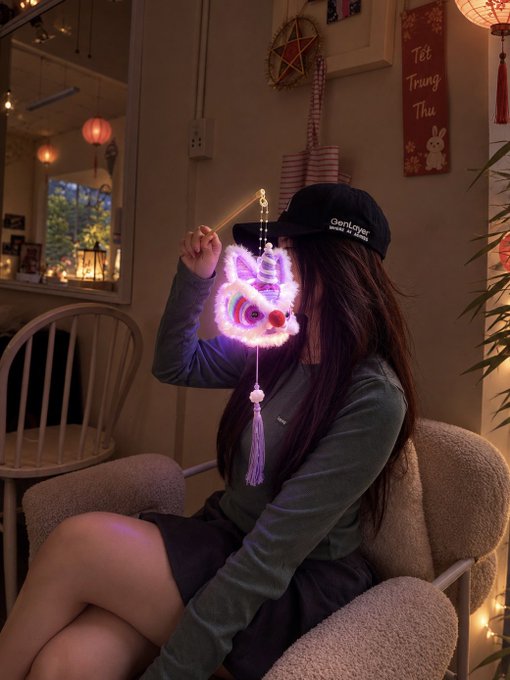 A person wearing a black cap and green top sits in a dimly lit room holding up a glowing LED cat lantern that illuminates their face.