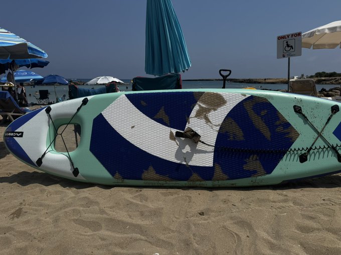 An inflatable stand-up paddleboard with a blue, white, and camouflage pattern labeled 'WBSUP' rests on a sandy beach near an umbrella and a handicapped-only sign.