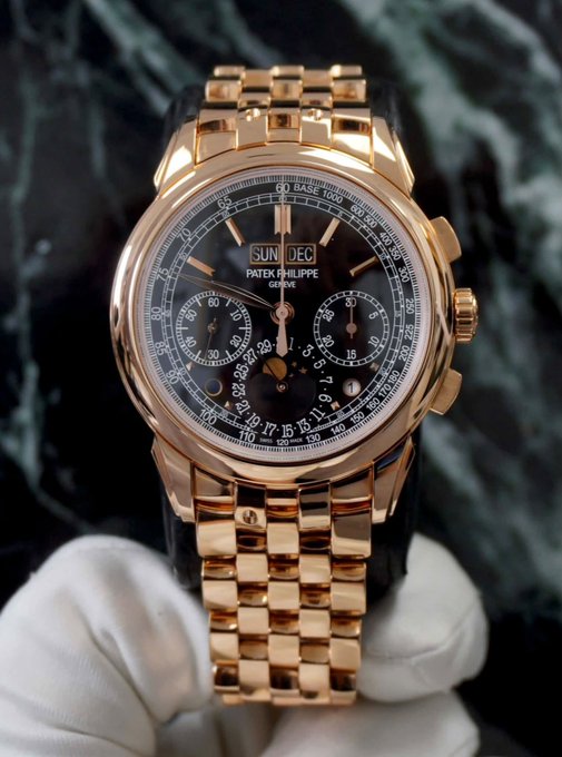 A luxury rose gold Patek Philippe watch with a black dial and chronograph subdials is held by white-gloved hands against a dark marble background.