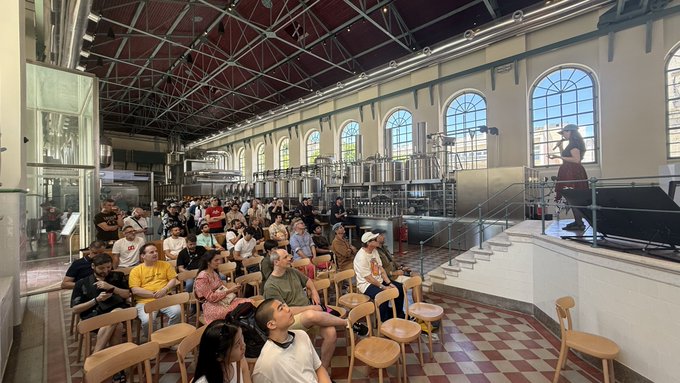 The image shows a large group of people sitting down and looking at something off-camera, possibly watching a speaker or an event taking place. The crowd consists of men and women who appear to be engaged in conversation with each other. There are chairs placed around them, indicating that they might have been arranged for this gathering. 

The image also contains the text "She's not the 'Queen of punks' she's not the 'princess of punks', she's the Joan of Arc of punks", which seems to be a reference or a humorous statement about the speaker or event they are watching. This adds an element of humor and lightheartedness to the scene, as it suggests that the people in the image might have been expecting a more serious or provocative speaker instead.