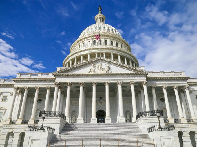 The image shows the U.S. Capitol Building with its iconic white dome and neoclassical columns, set against a bright blue sky with scattered clouds, an American flag flying atop. It serves as a symbolic backdrop for the post about the delayed CLARITY Act, visually representing the legislative body (Congress) where the bill's vote is now stalled, with declining odds of passage before 2027.