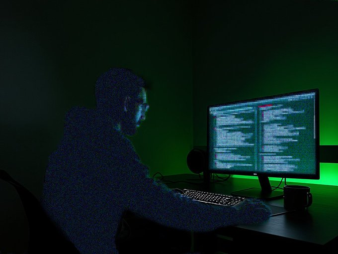 Silhouetted person working at a computer in a dark room illuminated by green backlighting, with code visible on the monitor.