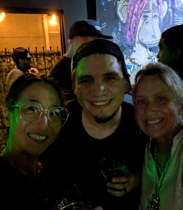 Three people posing together for a photo at what appears to be a bar or venue with graffiti art on the wall behind them.