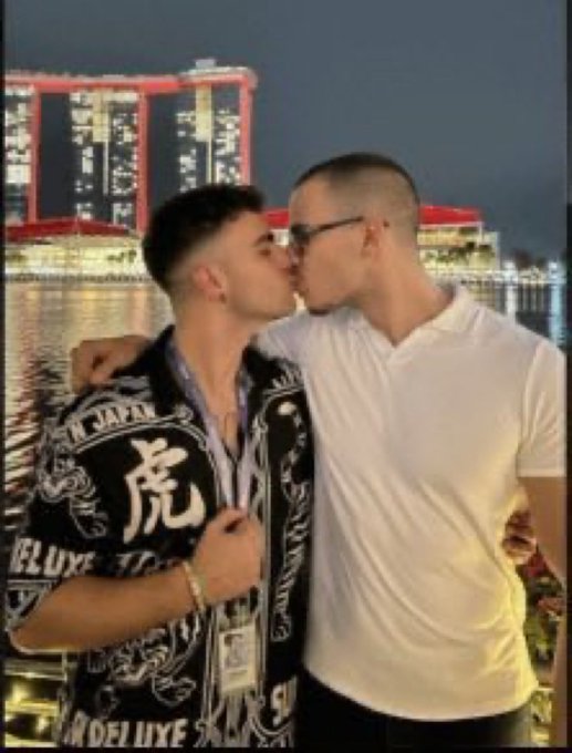 Two men share a kiss in front of Singapore's iconic Marina Bay Sands at night, with one wearing a black shirt featuring "JAPAN," "虎" (tiger), and "DELUXE" graphics, while the other wears a white polo shirt and glasses—appearing to capture a tender moment from a crypto conference experience.