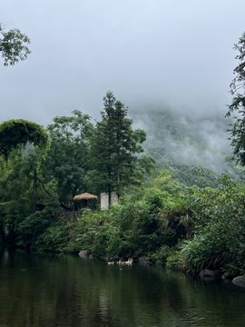 Mist drifts over lush forested hills behind a calm, mirror-like lake, where a small red pavilion peeks through the dense greenery—turning a simple snapshot into a cinematic, postcard-worthy scene straight out of a Chinese landscape painting.