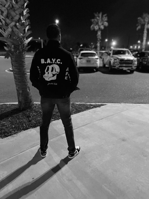 The image features a man standing alone on a sidewalk, wearing a black jacket with white text that says "BAYC Ape". The background shows several cars parked along the street, as well as palm trees nearby. There are also other people present in the scene, but they are not the main focus of the image.