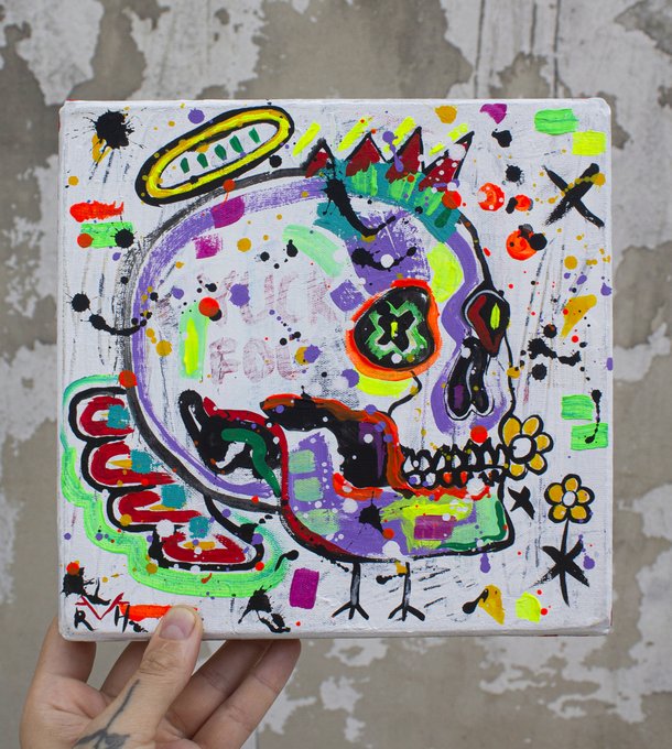 A hand holding a small colorful canvas painting of a decorated sugar skull with vibrant neon colors, flowers, a halo, and a cross, signed by the artist.