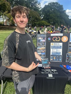 A smiling young man in a black t-shirt stands at an outdoor campus booth for "Blockchain at UMD," holding a card with information about "What is Blockchain?," "What is Web3?," and a Bitcoin logo, illustrating the post's mention of students from @blockchainatumd distributing flyers on the University of Maryland campus.