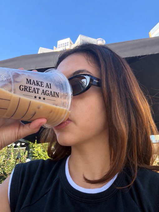 A woman wearing sunglasses drinks an iced coffee from a cup labeled 'MAKE AI GREAT AGAIN ★★★' against a clear blue sky.