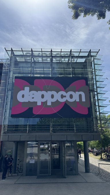 The image shows a large building with a glass facade, featuring a large sign that reads "dappcon". A group of people can be seen standing outside the building, possibly attending an event or gathering related to the dappcon conference. The scene is set against a backdrop of trees and a blue sky, creating a pleasant atmosphere for the attendees.

There are several individuals visible in the image, with some closer to the foreground and others further back near the glass facade of the building. A bicycle can also be seen parked nearby, possibly belonging to one of the attendees or staff members at the event.
