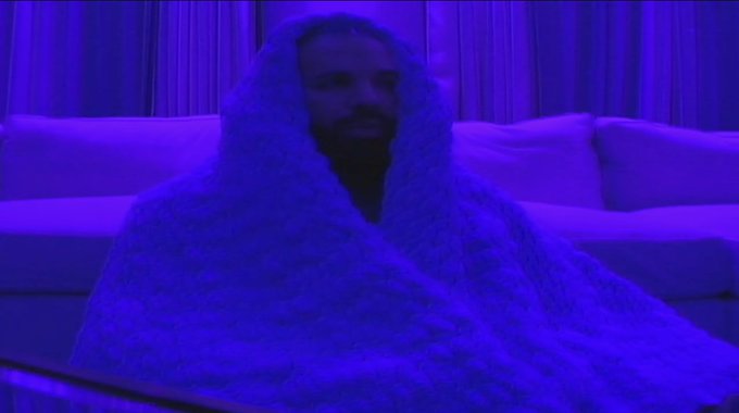 A man with a beard wrapped in a blanket, illuminated by purple lighting in what appears to be a bedroom or lounge setting.