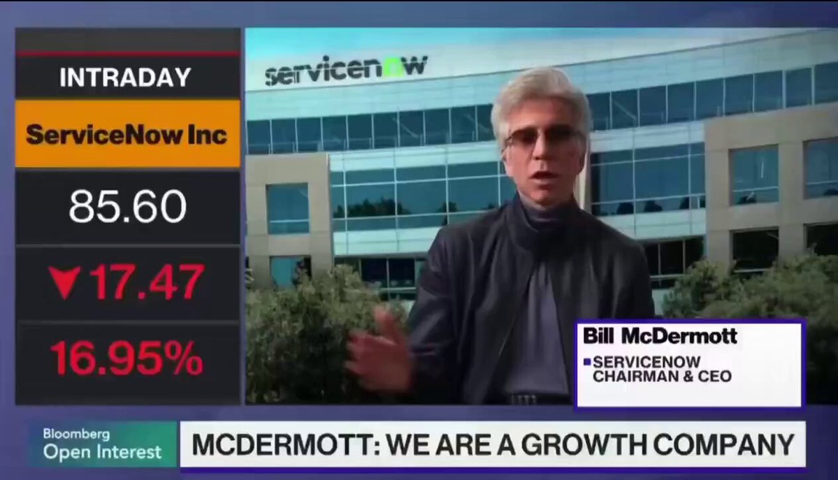 The image shows a news segment featuring a man talking about ServiceNow, Inc., which is a company that provides IT solutions for businesses. The man is wearing glasses and has short hair. He is standing in front of a large building with a glass facade, possibly an office or a store. The news segment also includes information about the stock market, as indicated by the text "Boomberg" at the bottom left corner.
