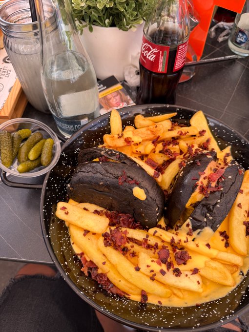 A bowl of fries topped with what appears to be black charcoal buns and bacon bits, served alongside a small dish of pickles and a bottle of Coca-Cola. The post highlights that the meal cost $13, paid via Tangem Pay, prompting the question of whether the unique presentation was worth the price.