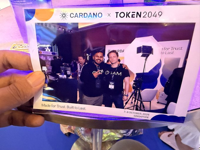 A hand holds a printed Polaroid showing them with another man at a 'CARDANO x TOKEN2049' booth, dated '7-8 OCTOBER 2025 SINGAPORE' with the slogan 'Made for Trust. Built to Last.'