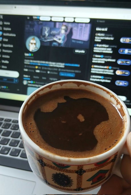 A hand holds a patterned ceramic mug of coffee in front of an open laptop displaying what appears to be a social media feed with blurred user interactions, fitting the "GM" (good morning) crypto Twitter greeting with a morning brew alongside timeline activity.