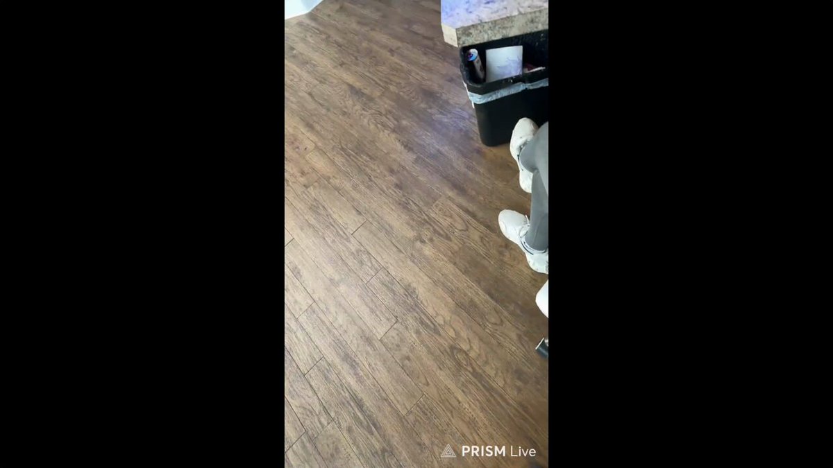 The image shows a person wearing white shoes standing on a hardwood floor, with their feet positioned near the bottom of the frame. The background features a black countertop that extends across the width of the photo. In the top left corner of the image, there is text that reads "this is what onboarding normies looks like new pumpfun bounty feature @Pumpfun @gorillacap".
