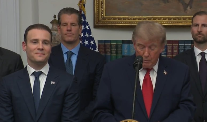 Donald Trump speaks at a podium while a smiling man in a suit stands to his left, wearing a pin that appears to read "HYPE".