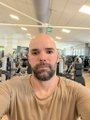 A low-resolution selfie of a bald, bearded man wearing a tan t-shirt in what appears to be a gym.