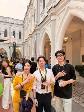 Three people pose for a photo at what appears to be an event in a courtyard with white archways; the woman on the left wears a yellow dress, while the two men wear lanyards.