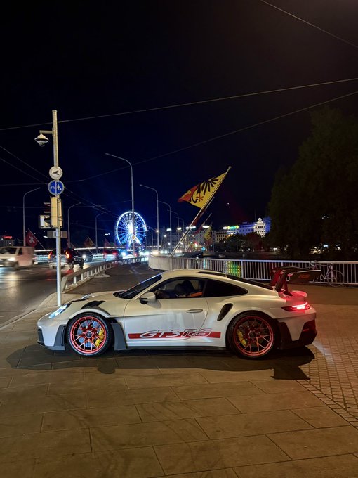 A white Porsche 911 GT3 RS with red wheels and red 'GT3 RS' side decals is parked at night on a city street, with a lit Ferris wheel and a flag visible in the background.