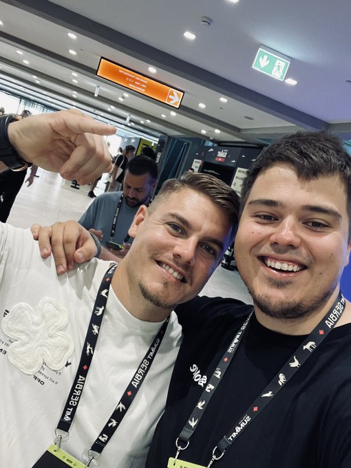A selfie of two smiling young men wearing lanyards in what appears to be a convention center or airport.