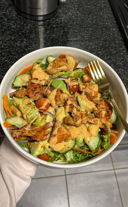 A fresh salad with grilled chicken, cucumbers, tomatoes, and carrots drizzled with a creamy orange dressing sits in a white bowl, paired with a gold fork on a tiled countertop. The post reflects a positive, grateful vibe, and the healthy meal visually echoes the "blessed" and energizing tone of the message.