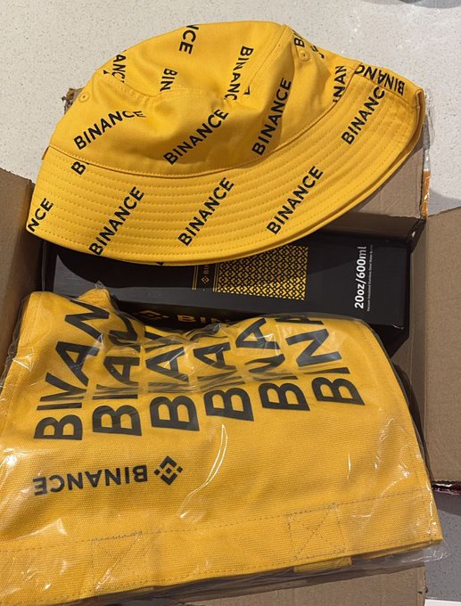 The image features a yellow bucket hat with black text that says "Binance Bucket Hat" and "Binance Binance". The hat appears to be made of plastic, as indicated by its shape and material. It's placed inside a cardboard box or a brown paper bag, which is sitting on top of a white surface like a table or countertop. There are also other items in the background that include a black book with gold lettering and a steel bottle.