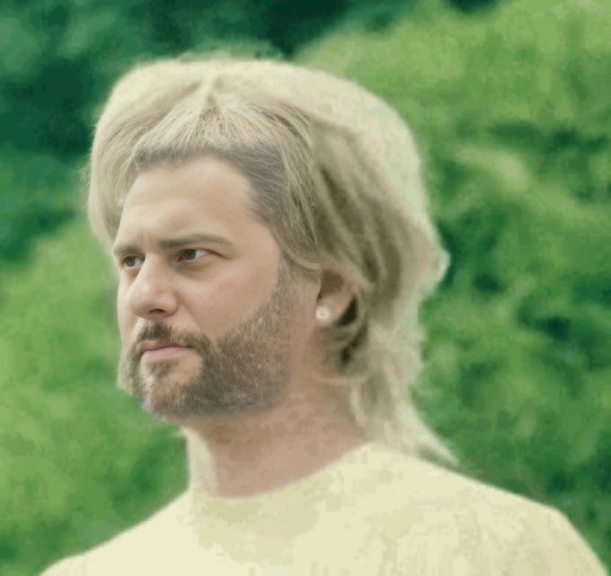 A portrait of a person with styled grey hair and a beard, wearing a light-colored top against a blurred green background.