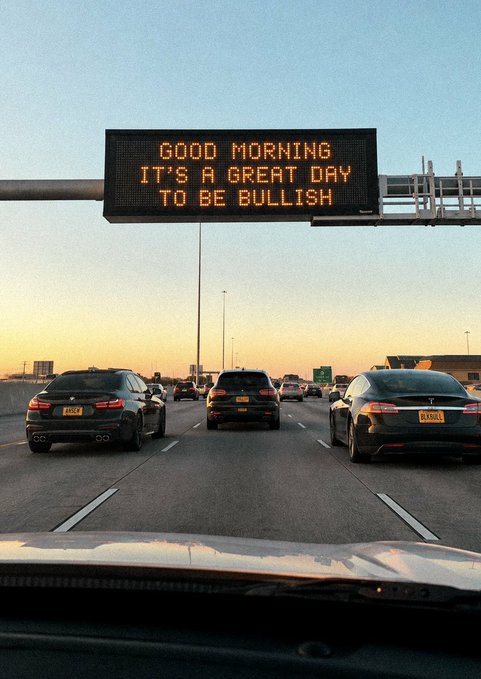 A highway overhead digital sign displays the message "GOOD MORNING IT'S A GREAT DAY TO BE BULLISH" in orange text above several cars driving during sunset.