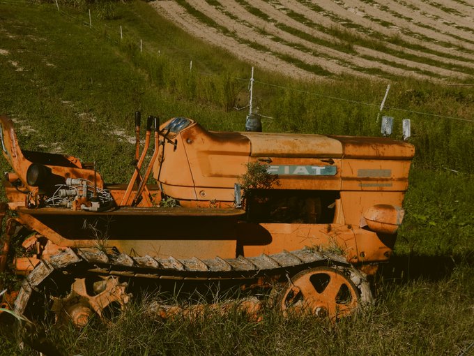 An abandoned orange Fiat tractor overgrown with grass in a field, set against a backdrop of plowed farmland rows.