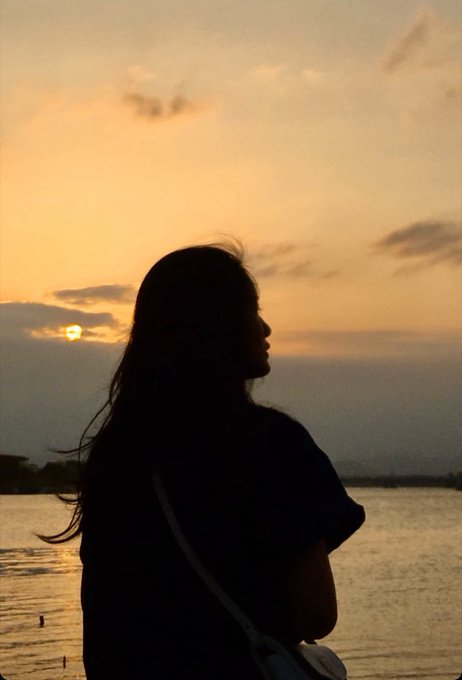 A silhouette of a person with long hair stands by the water, watching a golden sunset.