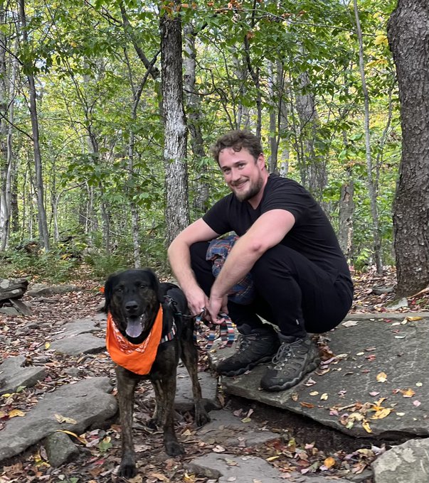 A man with a beard wearing a black t-shirt crouches on a rock in a forest next to a black dog wearing an orange bandana.