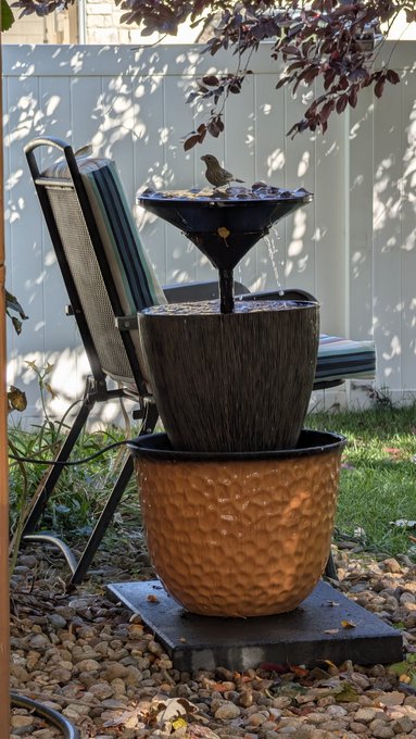 A decorative tiered water fountain sits in a backyard next to a folding chair, with a small bird perched on its top basin.