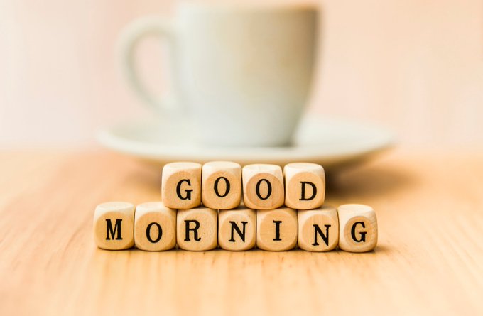 Wooden letter blocks spelling 'GOOD MORNING' arranged in front of a blurred white coffee cup and saucer on a wooden table.
