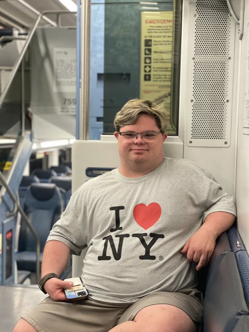 A young man wearing glasses and a gray 'I ❤ NY' t-shirt sits on a public transit train holding a transit card.