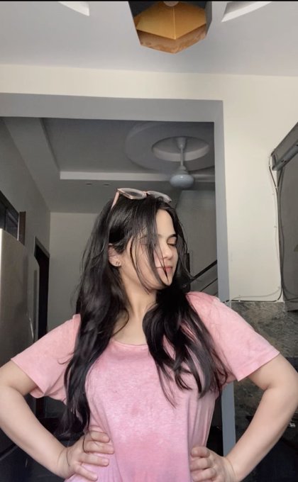 A young woman with long dark hair stands indoors wearing a pink t-shirt, with sunglasses perched on her head and her hands on her hips.