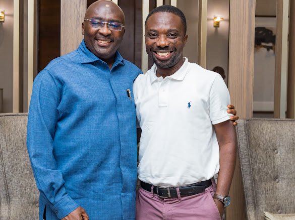 Two smiling men pose together for a photo; the older man wears a blue traditional shirt, and the younger man wears a white polo shirt with pink pants in a modern interior setting.