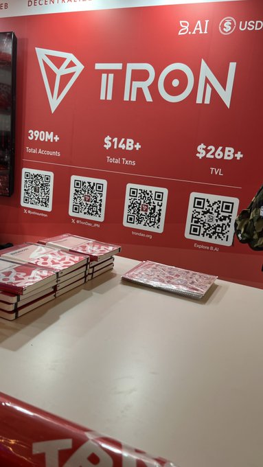 A red promotional booth for 'TRON' cryptocurrency displays statistics including '390M+ Total Accounts', '$14B+ Total Txns', and '$26B+ TVL', along with four QR codes and stacks of branded booklets on the counter.