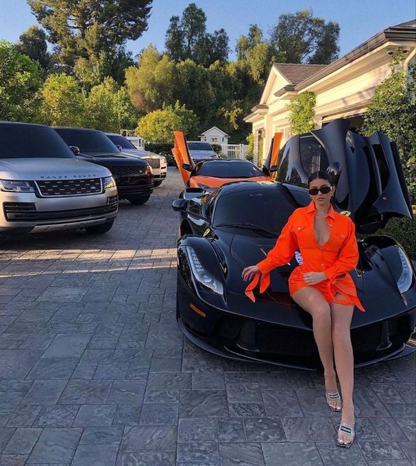 The image features a woman sitting next to an orange sports car with her legs crossed while wearing an orange dress. The car has its hood open, revealing its engine. In the background, there are other cars parked nearby. The caption of the image reads "Good night CT I heard @quipnetwork is launching in Q2 how true is that Anyways see you tomorrow, have a splendid night".