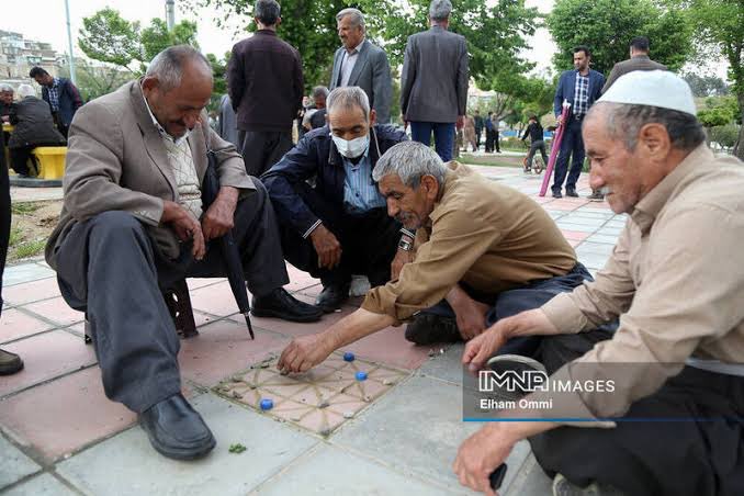 Older men gather outdoors playing a traditional tile-based game on a paved walkway; one man is wearing a face mask, and the image is credited 'IMNA IMAGES - Elham Ommi'.