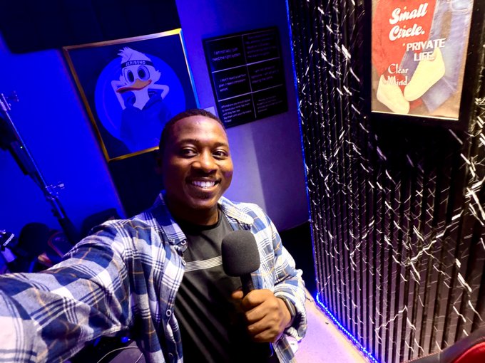 A cheerful man in a blue plaid shirt smiles while holding a microphone in a dimly lit studio featuring a framed Looney Tunes-style cartoon poster and a "Small Circle of Friends PRIVATE LIFE" movie poster on the wall, perfectly matching the upbeat "Good Morning" and "good day" greeting of his Saturday post.