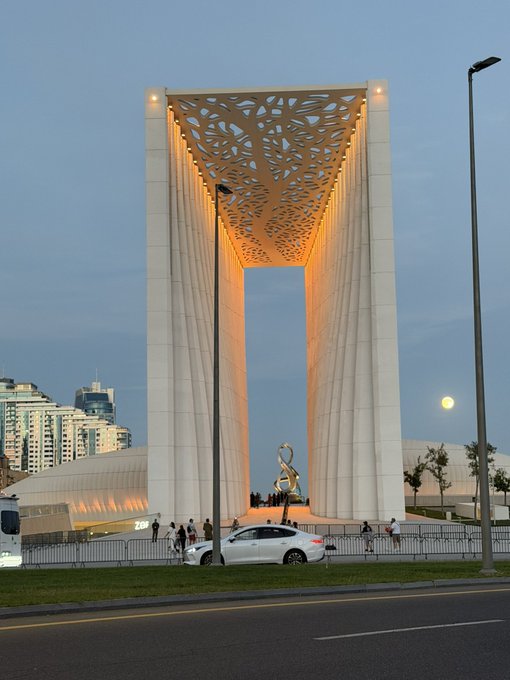 The image shows the iconic "Frame of Baku" monument illuminated at dusk, featuring its distinctive lattice-patterned ceiling and a statue visible in the central opening, with the moon rising in the background and a car passing in the foreground—perfectly capturing the architectural wonder mentioned in the caption praising Baku's beauty and inviting others to visit Azerbaijan.