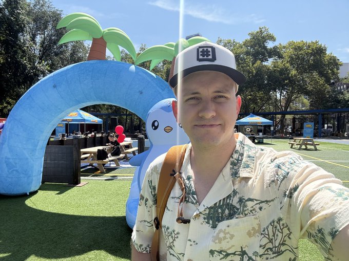 A man in a tropical print shirt and a black-and-white cap takes a selfie at a vibrant outdoor "Collector Park" event, with a large inflatable penguin and palm tree arch visible behind him on the artificial turf. The scene aligns with the post's mention of an OpenSea gathering, featuring branded tents and a playful crypto/NFT-themed atmosphere.