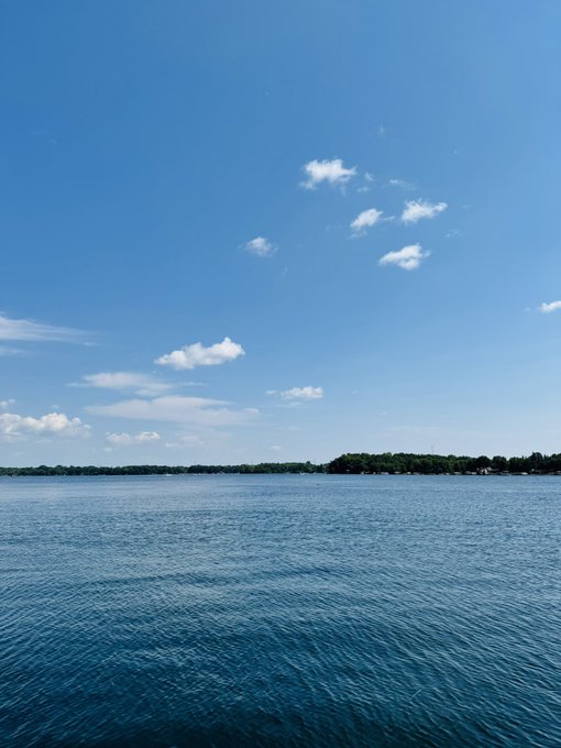 A serene lake stretches out under a vast blue sky dotted with wispy clouds, with a distant tree-lined shore on the horizon. The tranquil natural beauty pairs with the post's reverence for "God's creation," while the open water mirrors the boundless optimism in the accompanying Bitcoin and MSTR price predictions.
