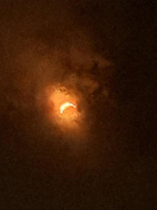 A close-up view of a solar eclipse with an orange-red glow, where clouds partially obscure the bright crescent of the sun.