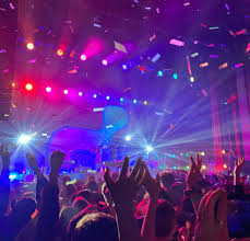 A vibrant concert scene with a crowd of silhouetted people raising their hands, colorful stage lights shining through, and confetti falling from the ceiling.