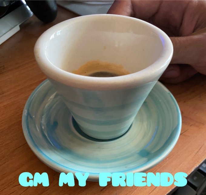 A close-up photo of a small ceramic coffee cup containing espresso, sitting on a matching blue and white saucer on a wooden table, with the teal text "GM MY FRIENDS" overlaid at the bottom.