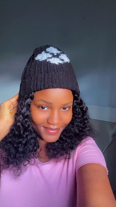 A woman with curly hair wearing a pink top and a black knit beanie featuring a white face/X logo takes a selfie.
