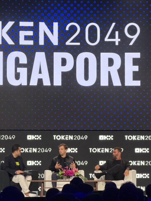 Three men sit on a stage having a panel discussion at the TOKEN2049 SINGAPORE conference, with the event's logo displayed prominently on the backdrop.