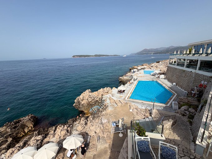 A scenic coastal view showing a cliffside resort with an infinity pool overlooking the Mediterranean Sea, featuring rocky shores, white umbrellas, and distant mountains under a clear blue sky.